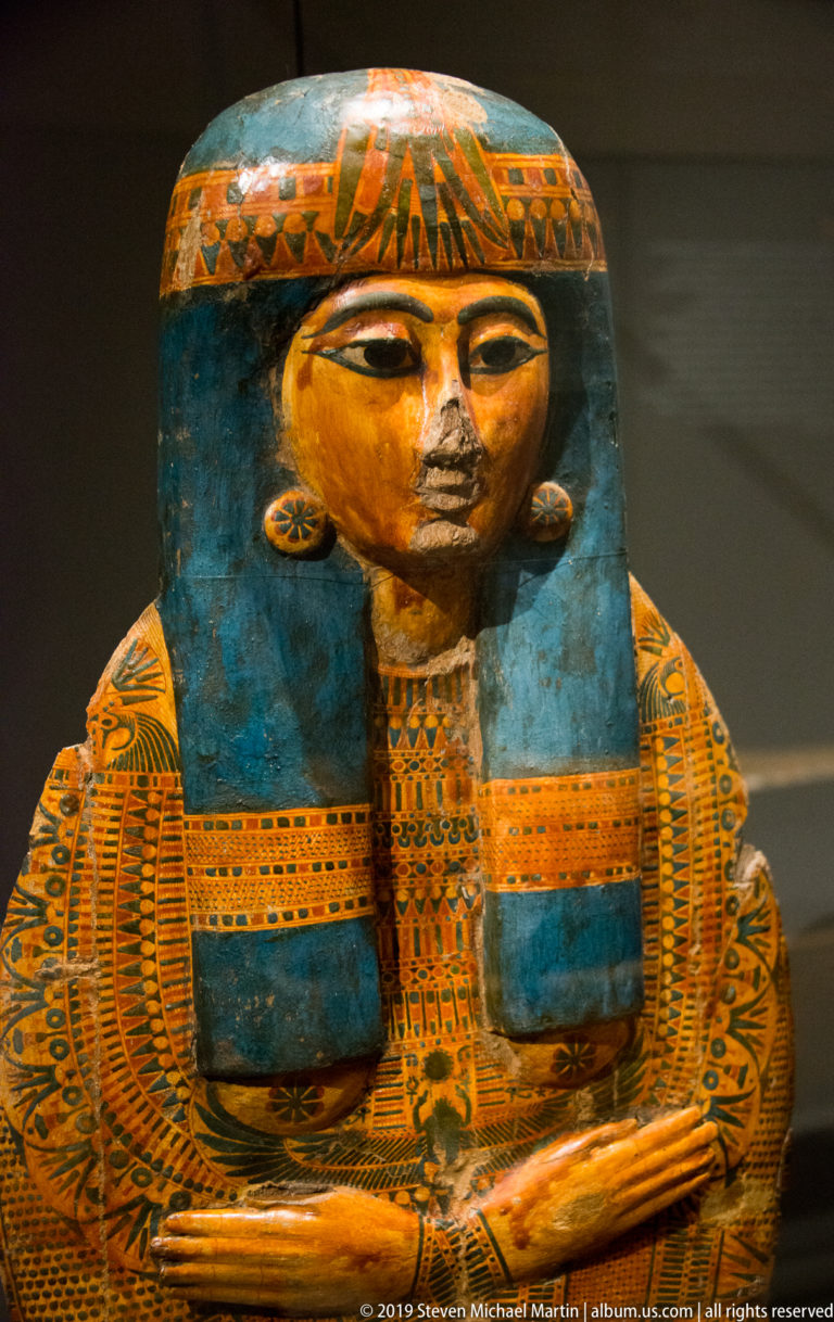 Frontal painted anthropoid coffin with gold-toned face, broad blue wig, large eyes, and dense orange-green chest decoration.