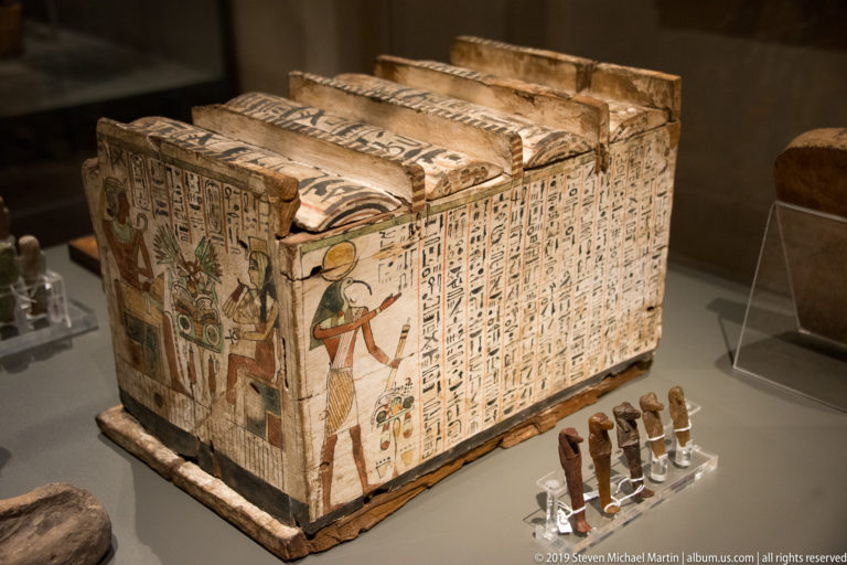 Rectangular Egyptian funerary chest covered with hieroglyphs, standing deities, and colored borders, displayed beside small figurines.