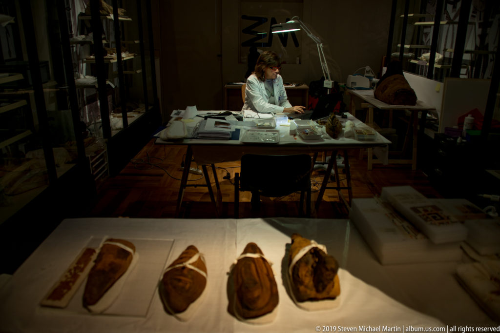 Conservator seated at a laboratory table with task lights and tools, while small wrapped or mummified objects lie in the foreground.