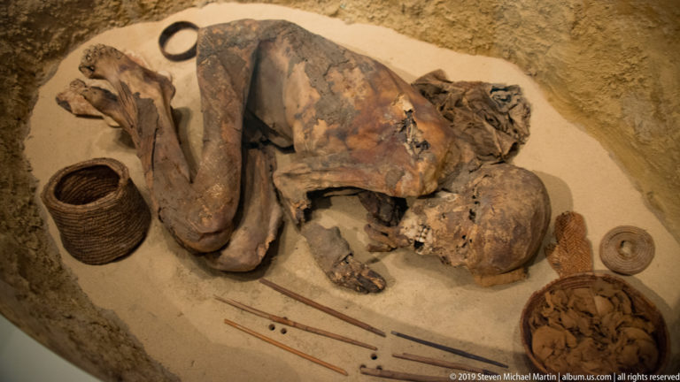 Contracted human burial in a shallow grave with knees drawn up, surrounded by baskets, vessels, and long narrow grave goods.