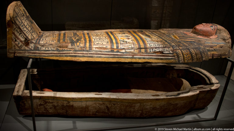 Open painted Egyptian anthropoid coffin with decorated lid raised above the lower case and a reddish wrapped form visible inside.