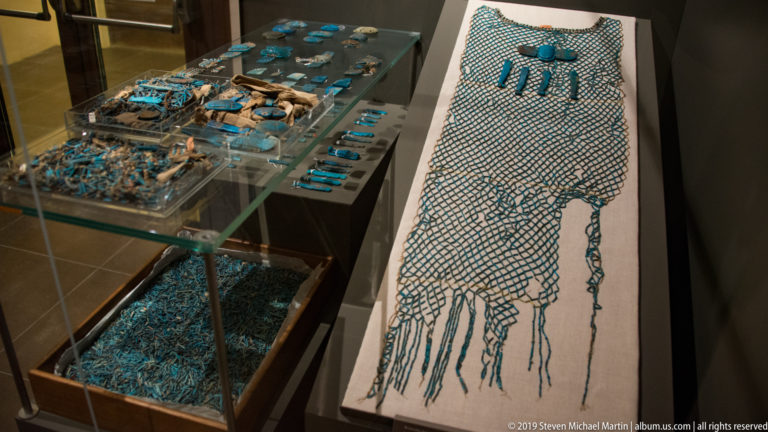 Blue-green Egyptian bead net displayed beside trays of small turquoise amulets and fragments in a glass case.