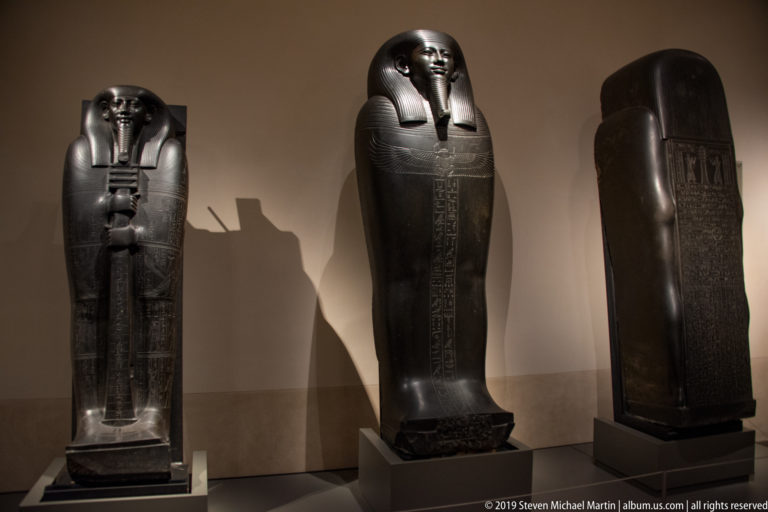 Three upright dark Egyptian anthropoid coffins displayed together, including inscribed and fully modeled examples.