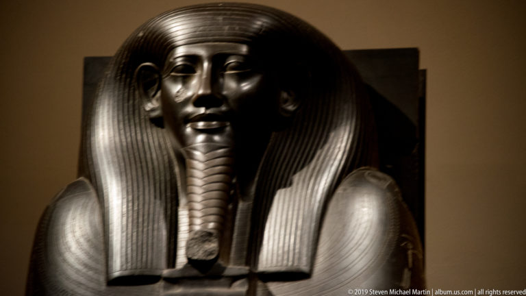 Polished dark anthropoid coffin face with striated wig, idealized features, and a long ceremonial beard.