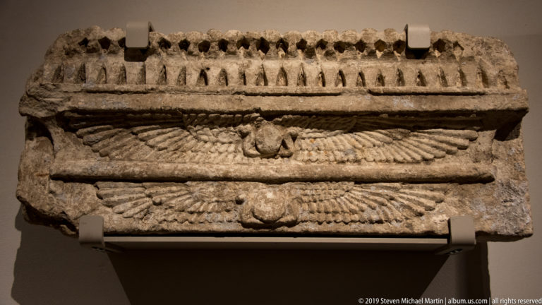 Rectangular carved stone fragment with two broad-winged motifs beneath a repeating geometric upper border.