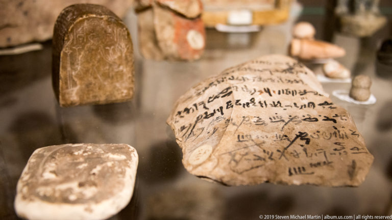 Large pale Egyptian ostracon covered with horizontal lines of dark cursive script, displayed with smaller fragments.