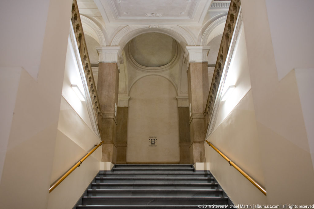 Symmetrical museum stairway with dark steps, brass-toned rails, pilasters, and a tall arched recess at the landing.