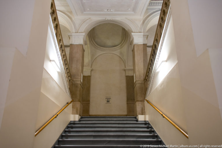 Upward view of a museum stairway with dark steps, brass-toned handrails, pale walls, and a tall arched landing.