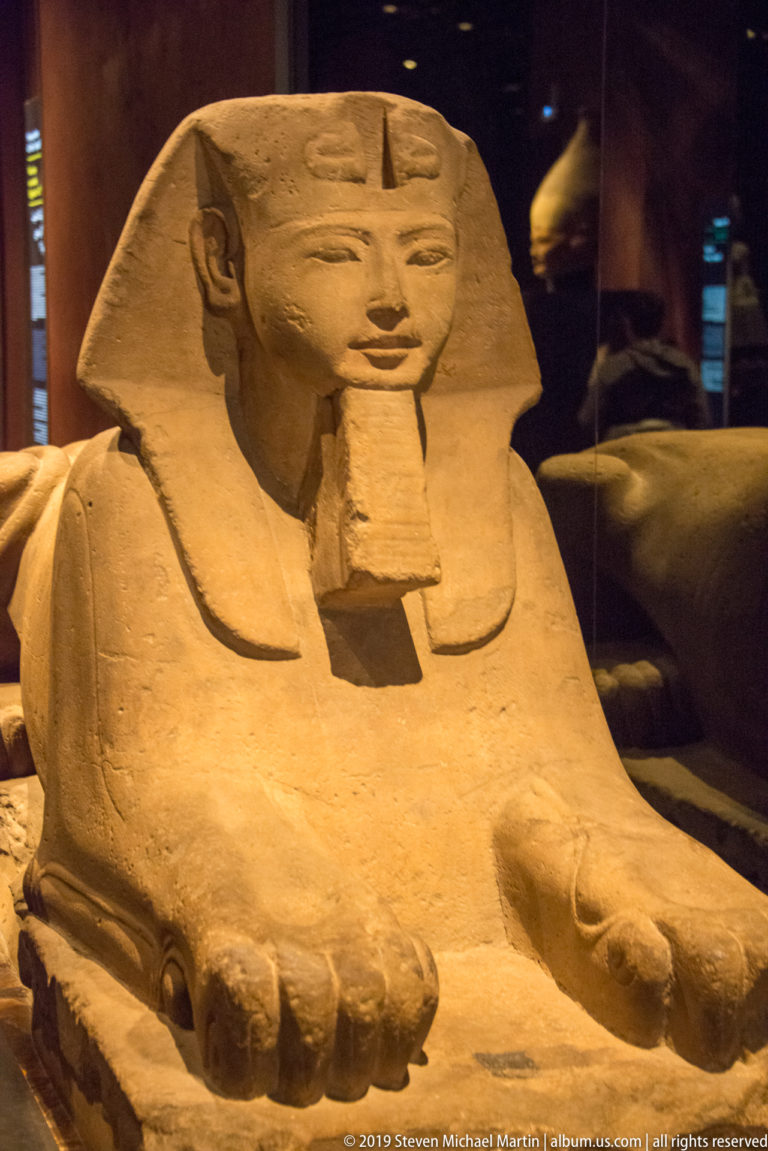 Frontal monumental sphinx with king's face, nemes headdress, beard, broad lion chest, and large forepaws.