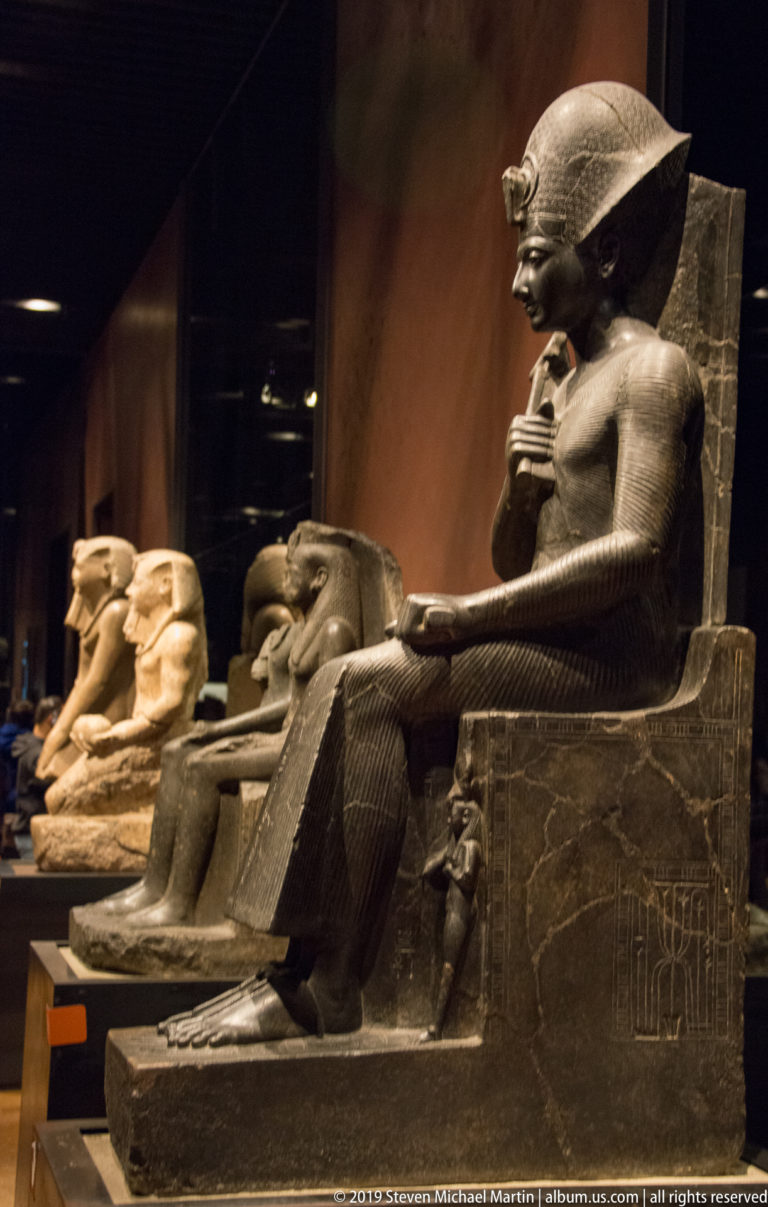 Profile of a seated Egyptian king on an inscribed throne, with additional statues receding in a dark museum gallery.