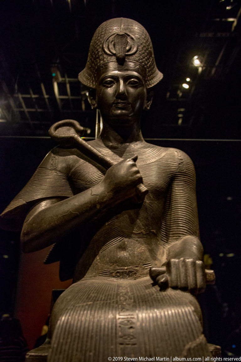 Dark stone seated Egyptian king holding a curved staff across his chest, with royal headdress and patterned garment.