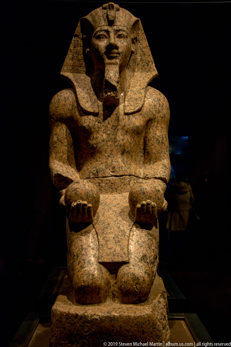 Kneeling granite statue of Amenhotep II wearing a nemes headdress and presenting two vessels in his open hands.