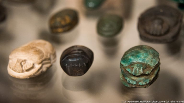 Small Egyptian scarab amulets and seals in pale, dark, and blue-green materials, some showing carved inscriptions on their undersides.