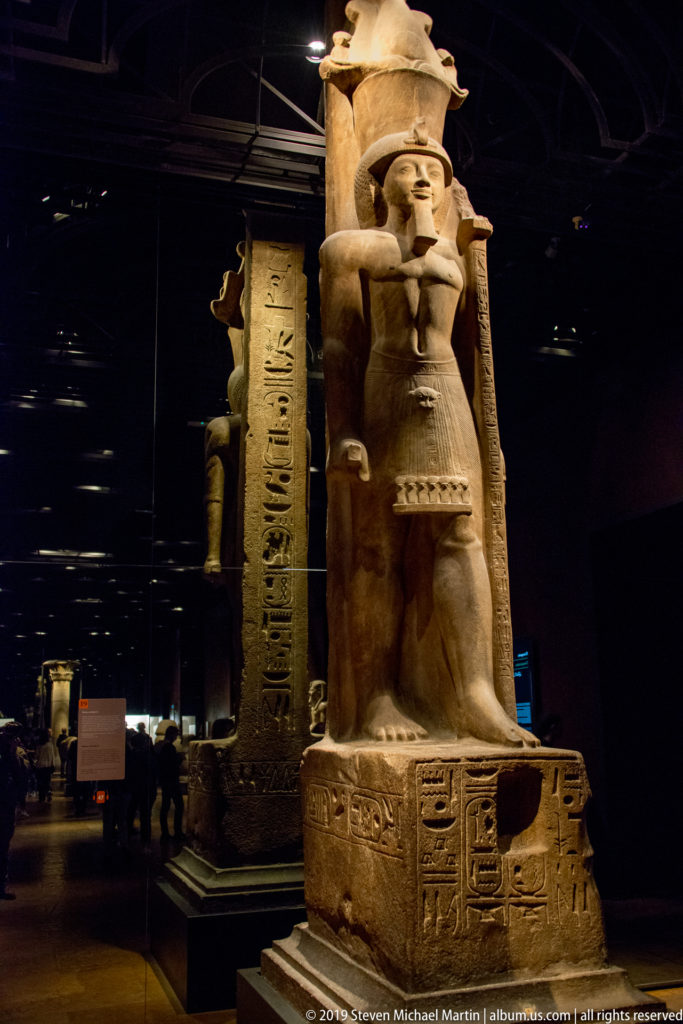 Standing Egyptian deity carved beside a tall hieroglyph-covered pillar and other sculptural elements in a dark gallery.
