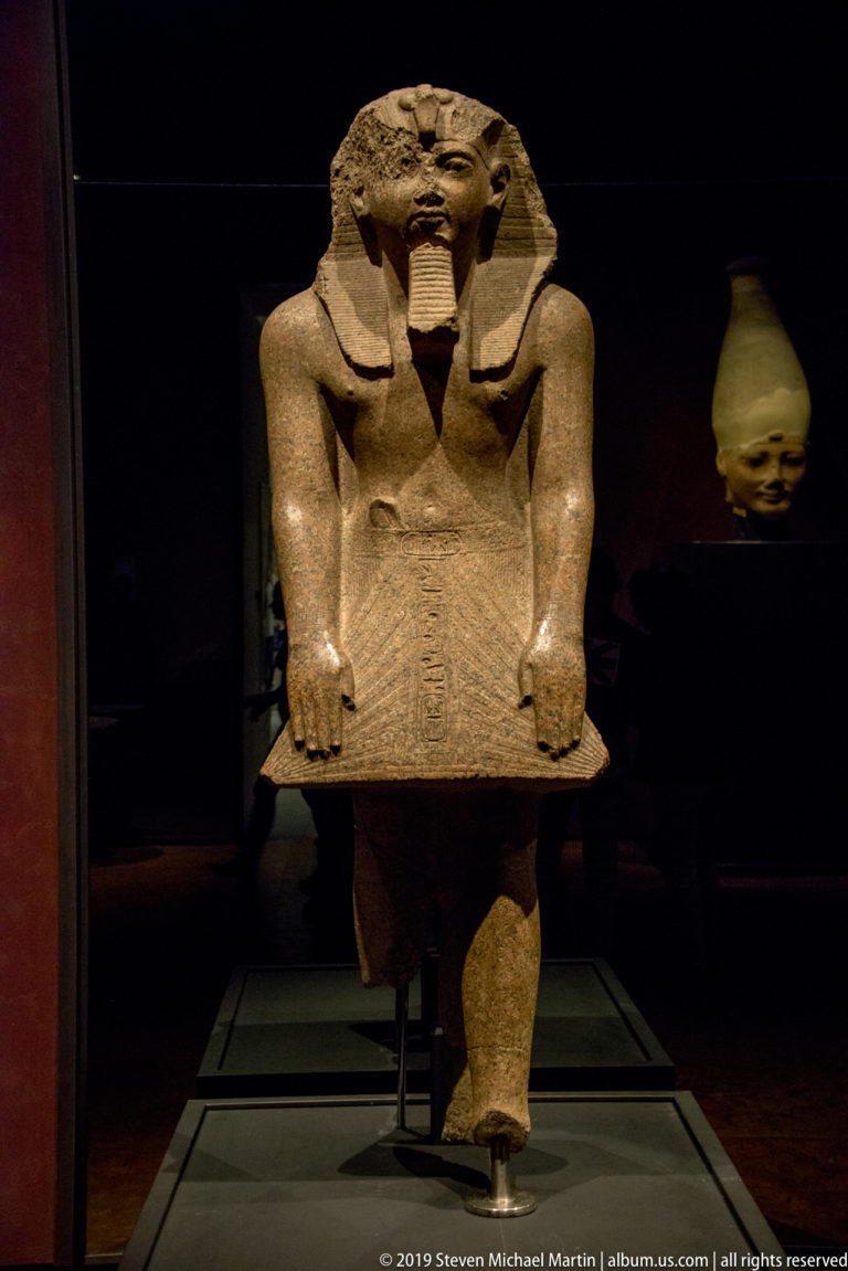 Fragmentary kneeling Egyptian royal statue wearing a striped headdress and beard, with a broad slab held or presented in front.