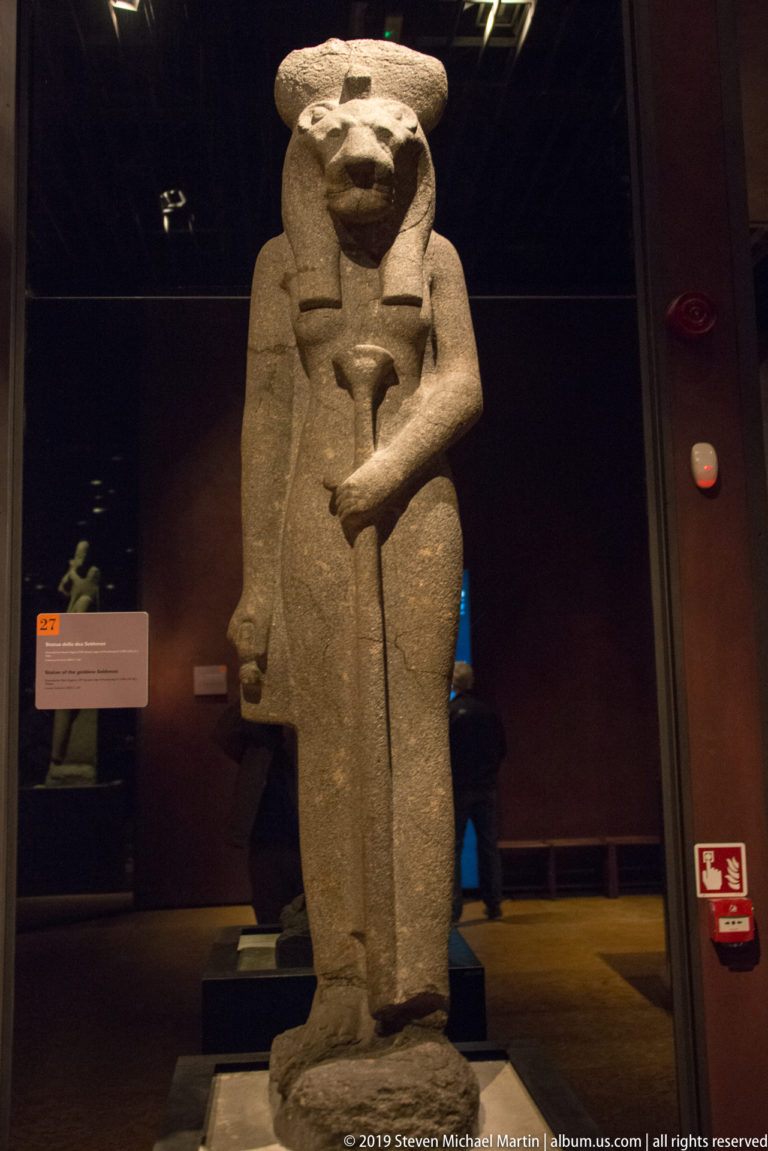 Standing granodiorite statue of Sekhmet with lioness head, solar disk, long dress, and vertical emblem held against her body.