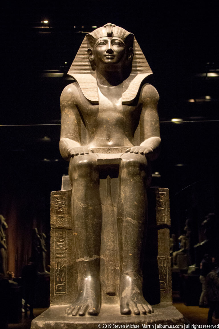 Monumental seated Egyptian king with striped headdress, hands on knees, and hieroglyphs carved down both sides of the throne.