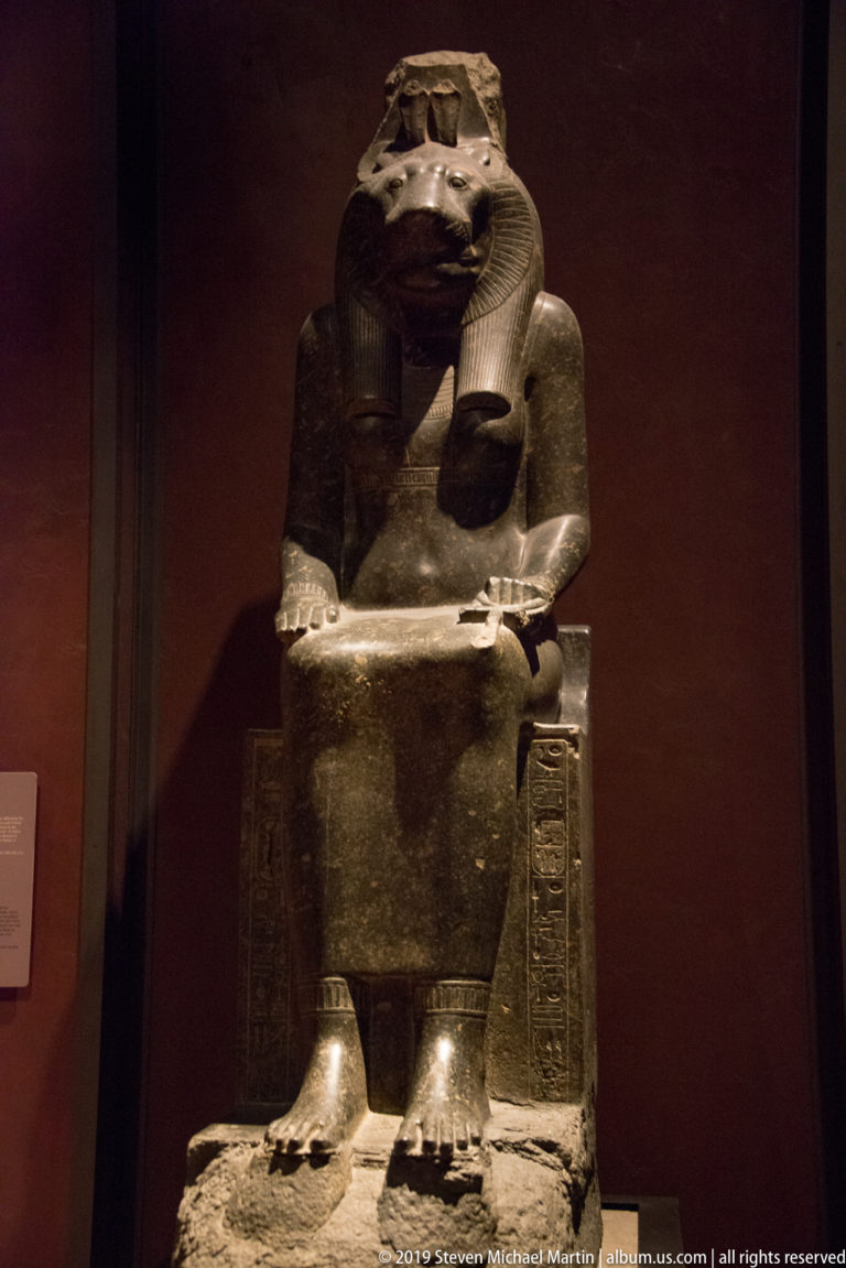 Dark stone seated pharaoh with striped headdress, hands on lap, and hieroglyphic inscription along the throne side.