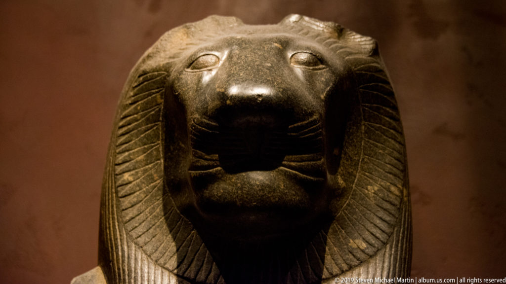 Frontal granodiorite statue of Sekhmet with lioness head, rounded ears, broad muzzle, and striated Egyptian wig.