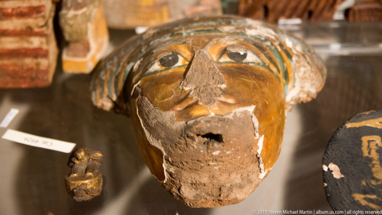 Close view of a painted Egyptian funerary face with large eyes, turquoise details, and extensive losses across the nose and mouth.