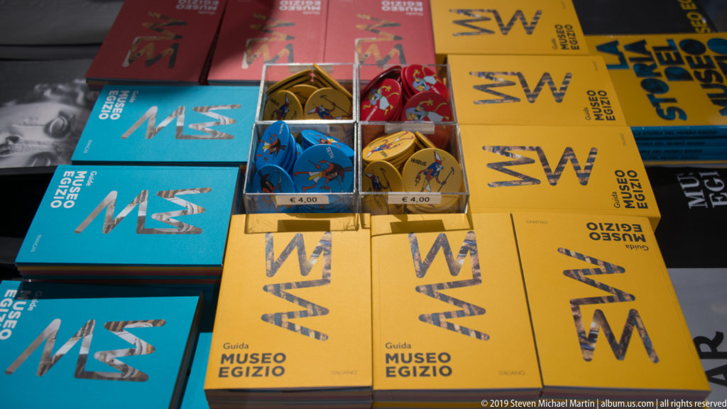 Overhead view of yellow and blue Museo Egizio guidebooks with small boxed souvenirs arranged on a shop table.