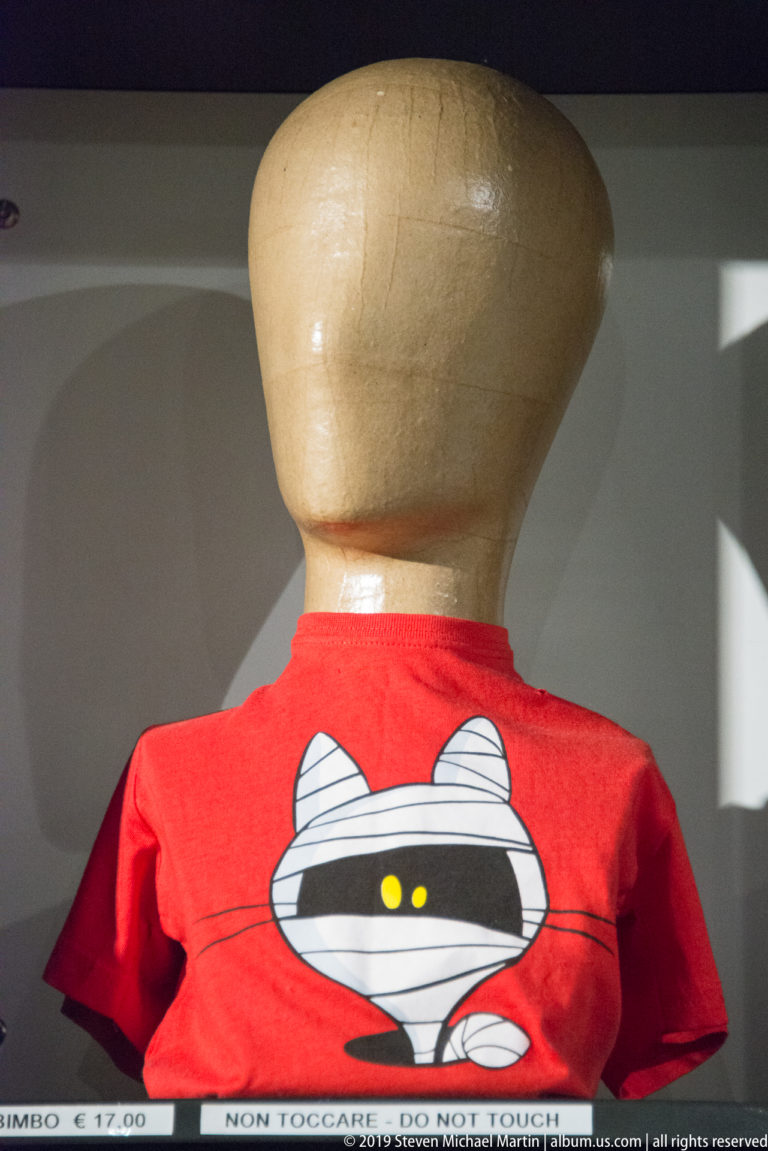 Red museum-shop T-shirt on a torso form, printed with a black-and-white cartoon mummy and yellow eyes.