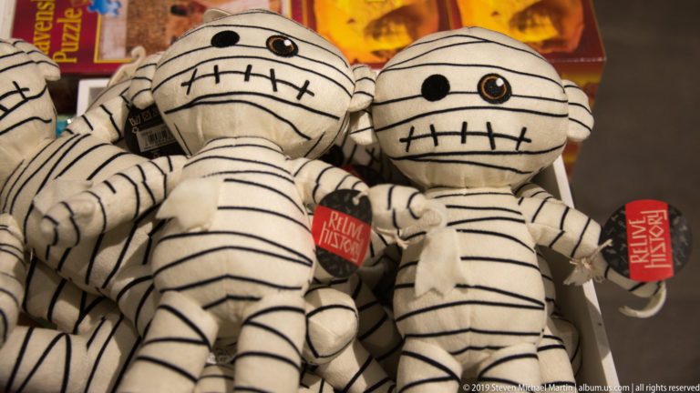 Pile of white plush mummy toys with black stitched wrappings, large eyes, and red retail tags in the museum shop.