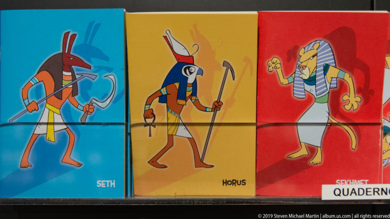 Colorful museum-shop notebooks with cartoon Egyptian deities, including covers labeled Seth and Horus.