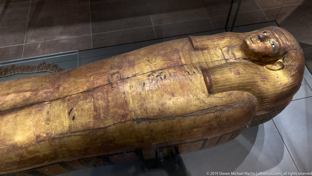 Full-length gilded Egyptian anthropoid coffin lying in a display case, with modeled face, headdress, and crossed forearms.