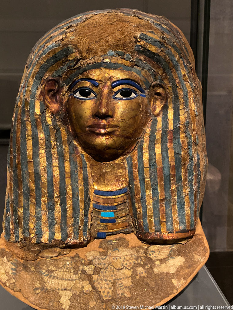 Frontal painted Egyptian coffin face with gold-toned skin, dark eyes, striped wig, and blue-green collar details.