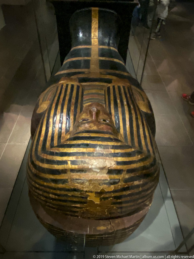 Rounded foot end of a black-and-gold striped Egyptian anthropoid coffin displayed horizontally in a museum case.