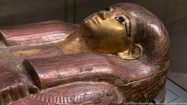 Angled close view of an Egyptian anthropoid coffin with glossy eyes, blue eye lines, striped wig, and crossed arms.