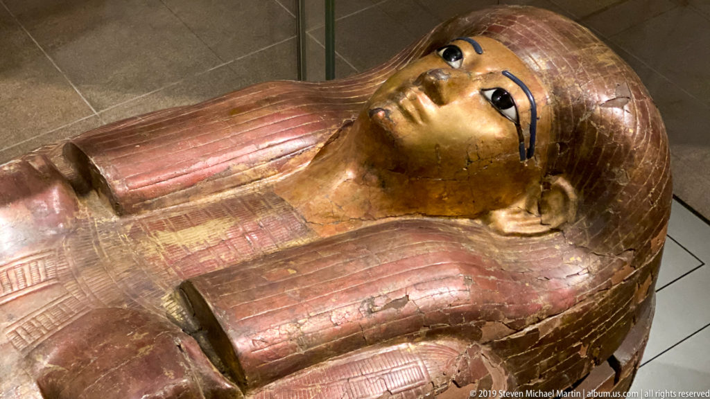 Oblique view of a gilded anthropoid Egyptian coffin with striped wig, crossed arms, and reddish-brown upper body.