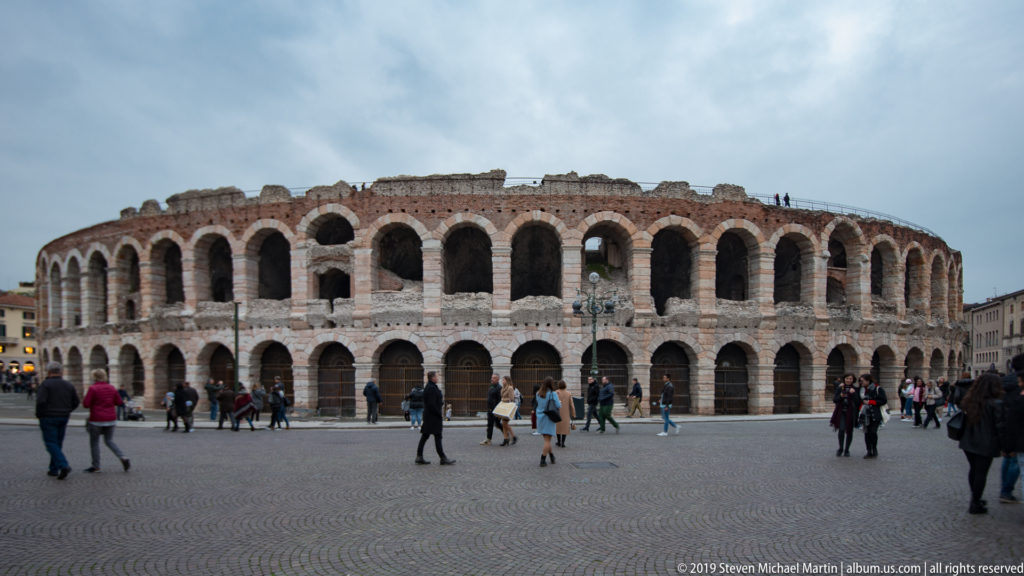 Arena di Verona seen across Piazza Bra, with two tiers of Roman arches and pedestrians on the cobbled square.