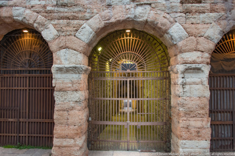 Close view of a gated Arena di Verona arch framed by alternating pale and reddish stone blocks.