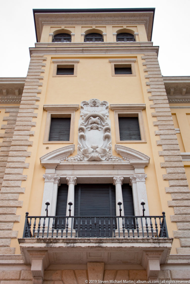 Upward view of a pale historic palazzo with balcony, paired columns, large sculpted mask, relief ornament, and banded masonry.
