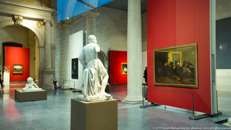 Castelvecchio Museum gallery with stone columns, sculptures on pedestals, paintings, and deep red display walls.