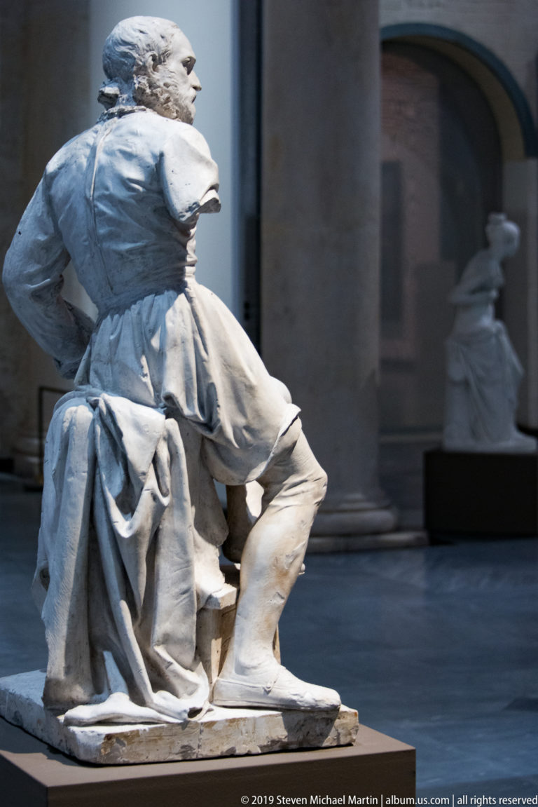 Seated pale stone sculpture with bent knee, heavy drapery, and turned head displayed inside Castelvecchio Museum.
