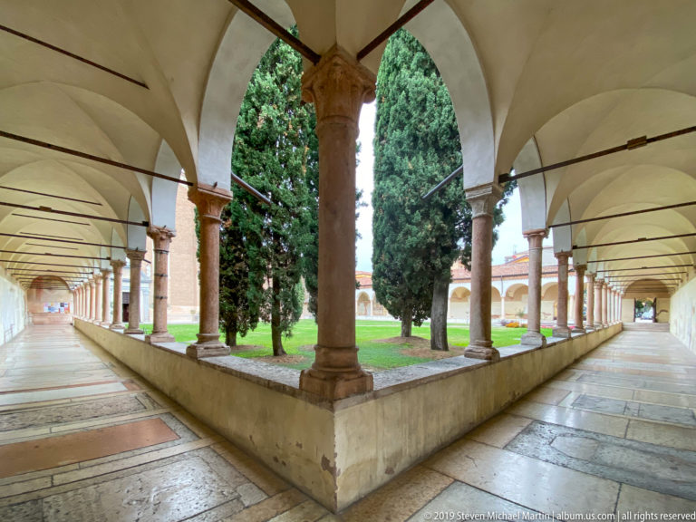 Corner of San Zeno cloister with intersecting arcades, stone columns, vaulted walks, paving, and tall cypress trees.