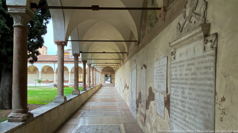 Long cloister corridor at San Zeno with arches, stone columns, wall-mounted inscribed slabs, and lawn at left.