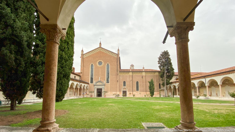 San Zeno cloister with stone arcades surrounding a lawn and the church façade centered beyond.