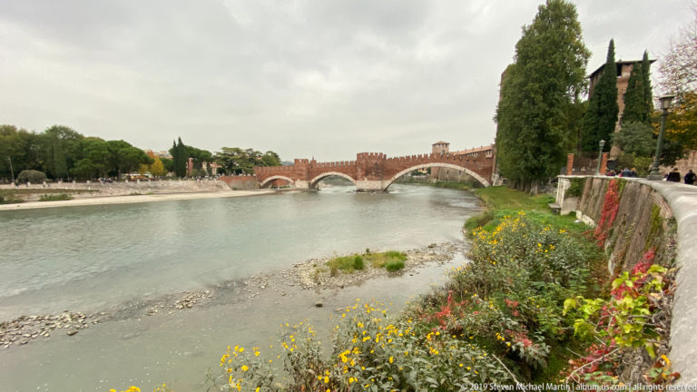 Ponte Scaligero crossing the Adige with pointed brick arches, crenellated parapets, and Castelvecchio fortifications beyond.