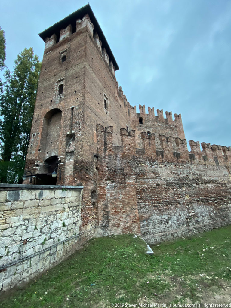 Close view of a square Castelvecchio tower joined to a crenellated brick wall above a stone base.