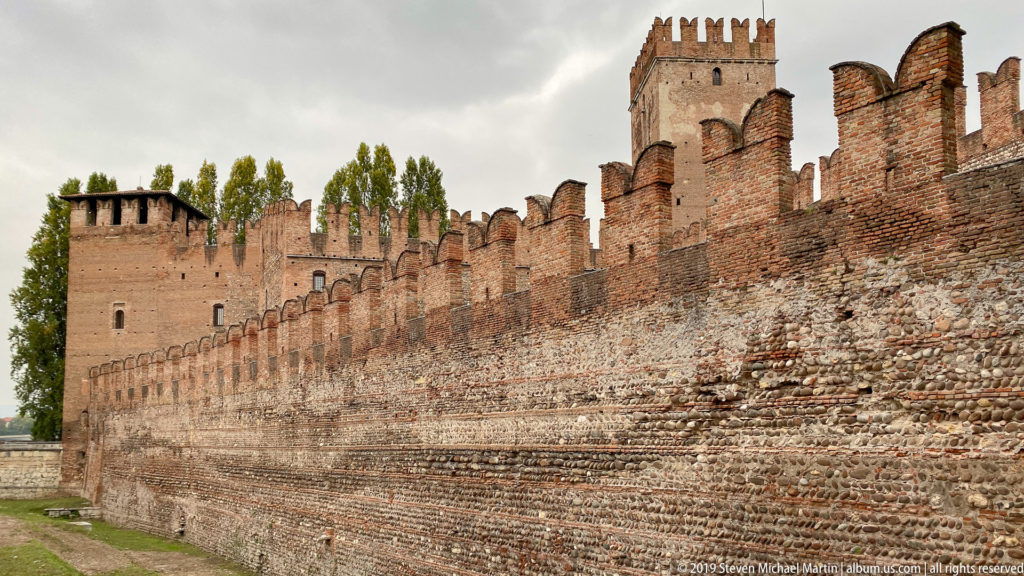 Long crenellated brick wall and towers of Castelvecchio seen obliquely beneath an overcast sky.
