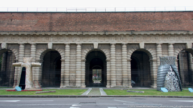 Two contemporary sculptures displayed on the lawn before the long rusticated arcade of Porta Nuova.