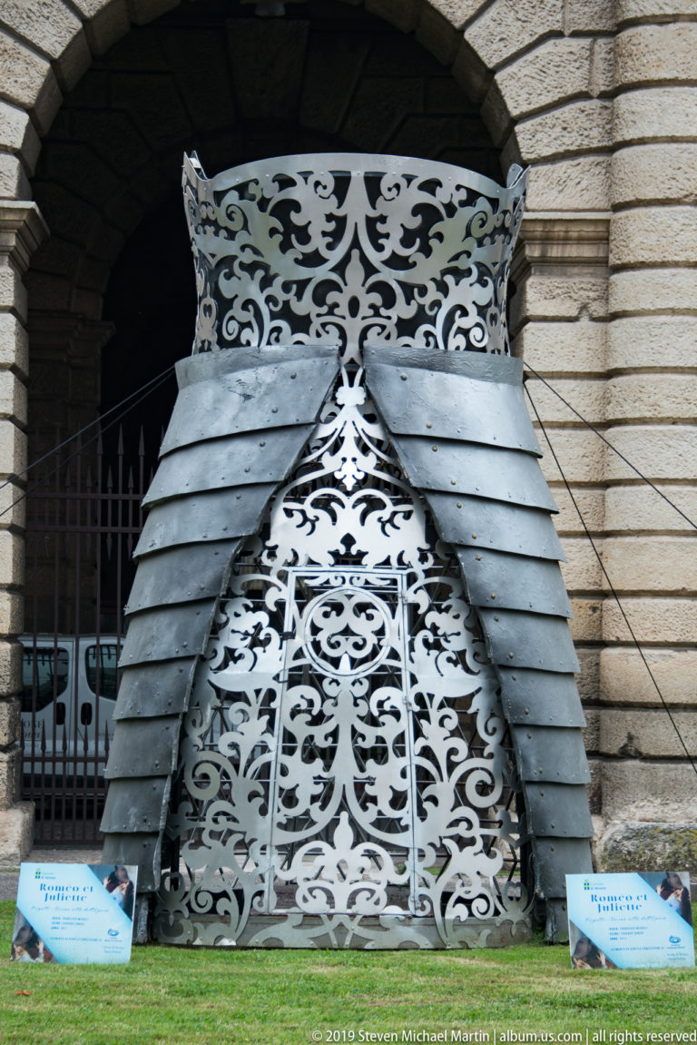 Tall pierced-metal sculpture with white scrollwork and layered gray plates standing beneath an arcade of Porta Nuova.
