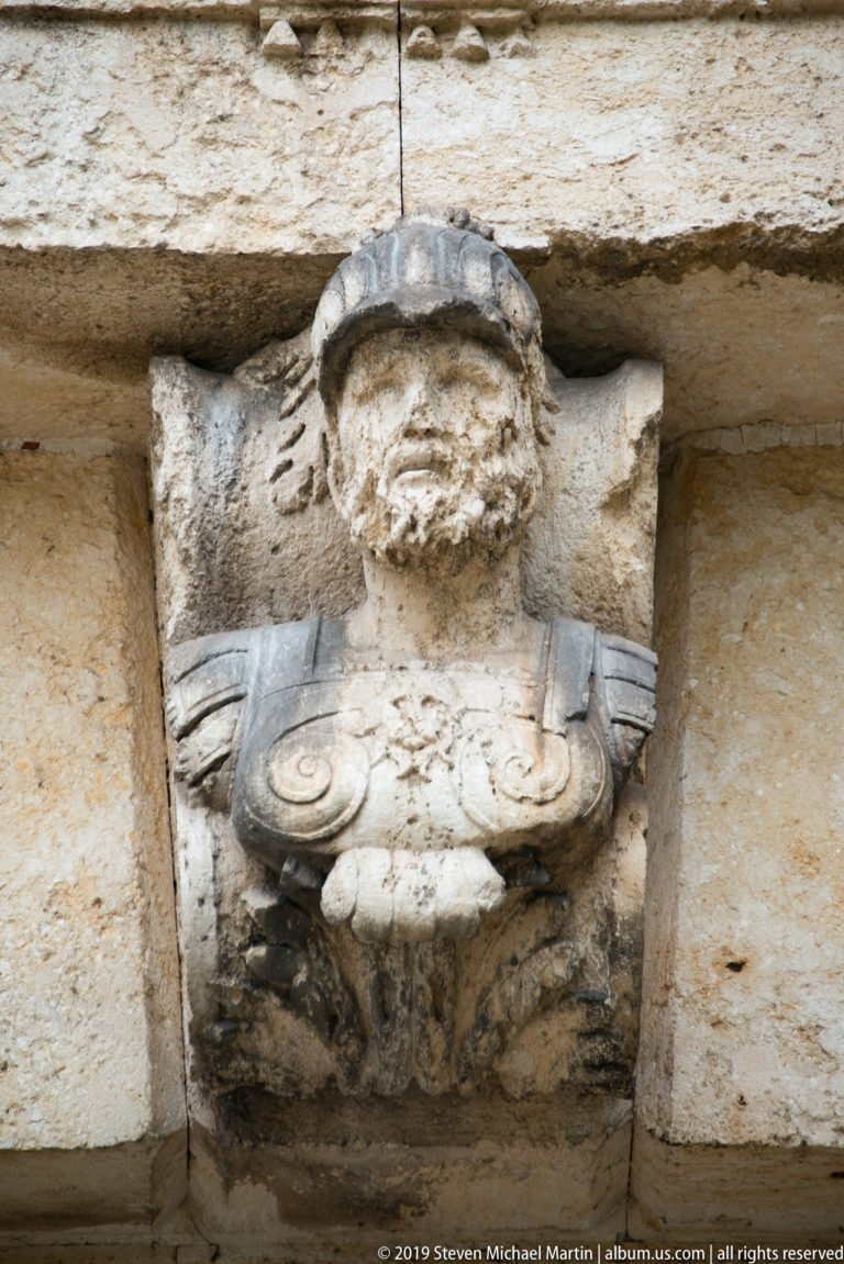 Weathered projecting stone head with heavy facial features and carved supporting forms on Porta Nuova.