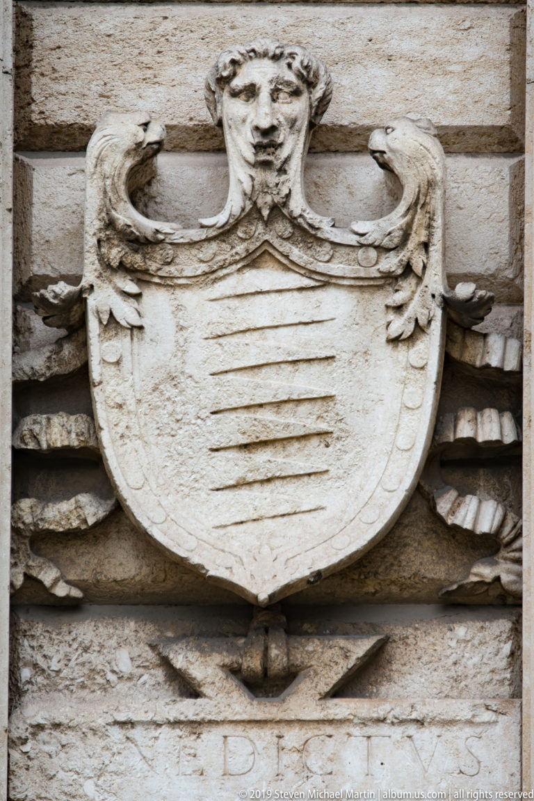 Weathered carved shield with horizontal bars, curling volutes, and a sculpted head on the stone façade of Porta Nuova.