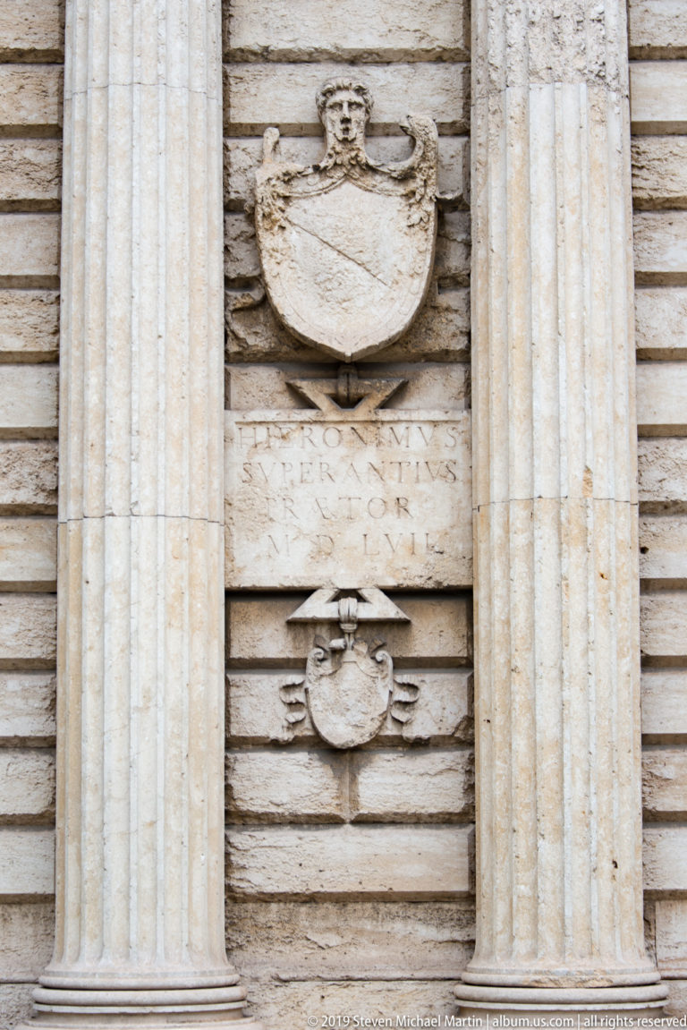 Two carved heraldic shields and a Latin inscription between fluted columns on the rusticated façade of Porta Nuova.