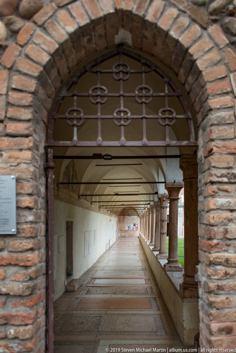 Long vaulted corridor seen through a brick arch, with wall panels at left and a courtyard arcade opening at right.